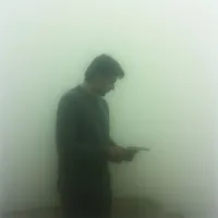 A man stands alone, looking down at something he holds in his hands. He’s enveloped by a thick, pale fog that obscures everything beyond him. The overall tone is somber and isolated, with the muted colors adding to the feeling of detachment.